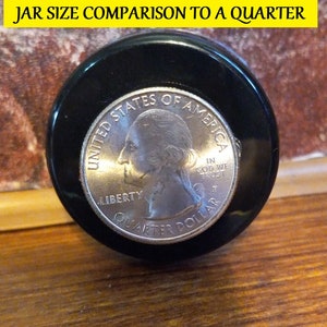 May include: A close-up of a US quarter coin, showing the head of George Washington, with the words "UNITED STATES OF AMERICA", "LIBERTY", "IN GOD WE TRUST", and "QUARTER DOLLAR" inscribed around the edge. The coin is sitting on top of a black jar lid.