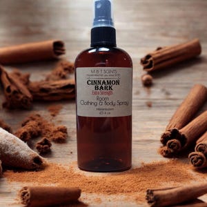 May include: A brown glass bottle with a spray nozzle, labeled "Cinnamon Bark Extra Strength Room & Body Spray" by M&T Scents. The bottle is surrounded by cinnamon sticks and ground cinnamon.