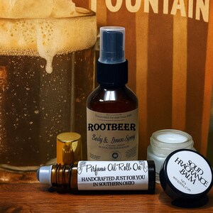May include: A collection of body care products including a brown glass bottle of Rootbeer Body & Linen Spray with a black pump, a small amber glass perfume oil roll-on, and a round black container of Solid Fragrance Balm with a white lid.