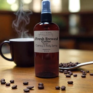 May include: A brown spray bottle labeled "Fresh Brewed Coffee Room Clothing & Body Spray" sits on a wooden surface. A dark brown coffee mug with steam rising from it is to the left. Coffee beans are scattered around the bottle and in a spoon.
