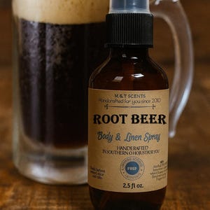 May include: A brown glass bottle of "ROOT BEER Body & Linen Spray" with a black spray nozzle. The bottle is in front of a beer mug filled with dark liquid and foam. The label reads "Handcrafted in Southern Ohio".