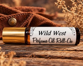 May include: A small amber glass perfume oil roll-on bottle with a gold-colored cap. The white label features black decorative flourishes and the printed text Wild West Perfume Oil Roll-On in a combination of serif and script fonts.