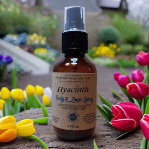 May include: A brown glass bottle with a black spray nozzle and a brown label. The label reads "M&T Scents Handcrafted for you since 2013 Hyacinth Body & Linen Spray HANDCRAFTED JUST FOR YOU IN SOUTHERN OHIO PARABEN FREE SULFATE FREE 2.5 fl oz."