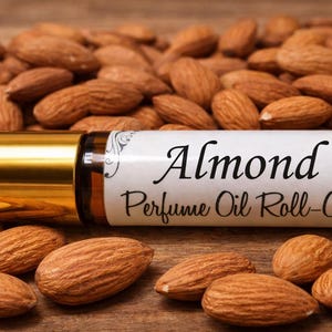 May include: A roll-on perfume oil bottle with a gold cap and a label that reads "Almond Perfume Oil Roll-On." The bottle is surrounded by a pile of almonds, creating a natural and aromatic theme.