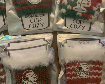 May include: Four handmade Christmas-themed cup cozies in individual clear plastic bags. The cozies are crocheted in red, green, and white with various holiday-themed appliques. The bags have a label that reads "handmade cup cozy" and "Clips By Coll".