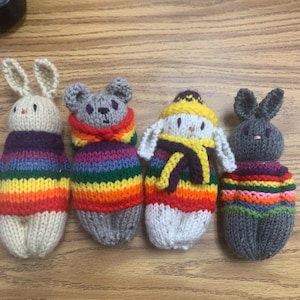 May include: Four knitted stuffed animals, each wearing a colorful striped sweater. The animals are a white bunny, a gray bear, a white snowman, and a gray bunny.