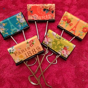 May include: Four binder clips with decorative paper covers. The covers feature floral patterns and the text "Paris 1889".