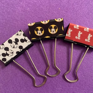 May include: Three silver binder clips decorated with different Mickey Mouse patterns. The first clip has a black background with yellow Mickey Mouse heads. The second clip has a white background with black Mickey Mouse heads and black polka dots. The third clip has a red background with white Mickey Mouse heads.