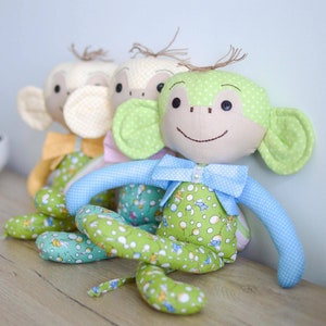 May include: Four handmade monkey plush toys in a row, each with a unique color scheme. The monkeys have button eyes, stitched smiles, and tufts of brown hair. They wear bow ties and patterned outfits in shades of green, yellow, and blue.
