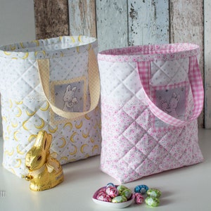 Może przedstawiać: Dwie materiałowe torby z pikowanymi wzorami. Jedna torba jest żółta z wzorem półksiężyca, a druga różowa z kwiatowym wzorem. Obie torby mają małą kieszeń z wzorem królika. Torby znajdują się na białym tle z czekoladowymi jajkami.