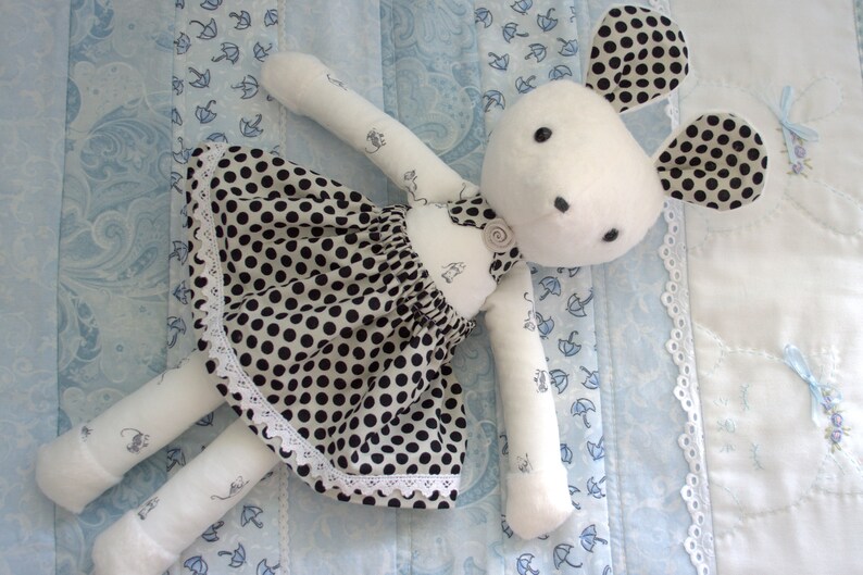 May include: A white stuffed toy rabbit wearing a black and white polka dot dress with white lace trim. The rabbit has large ears with black polka dots and a white face with black eyes.