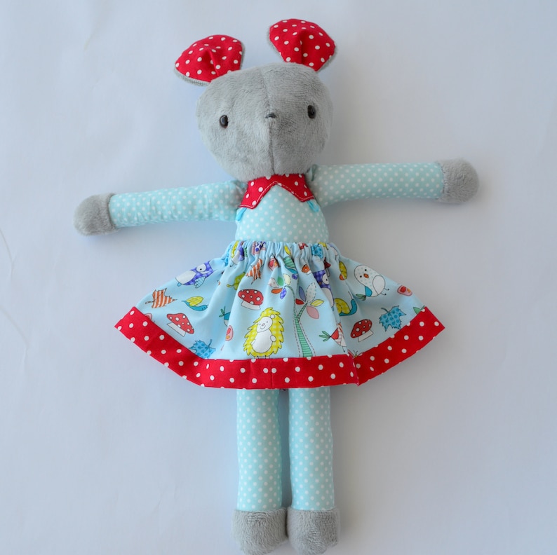 May include: A grey plush toy mouse wearing a blue dress with white polka dots and a red and white trim. The dress has a colourful print of birds, mushrooms, and hedgehogs. The mouse has red ears with white polka dots.