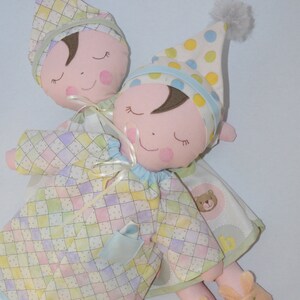 May include: Two plush dolls wearing matching pajamas with a checkered pattern in shades of yellow, pink, and blue. The dolls have embroidered eyes and smiles and are wearing matching hats with polka dots.