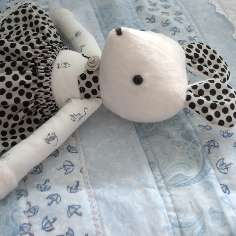 May include: A white plush toy mouse wearing a black and white polka dot dress. The mouse has black eyes and a black nose. The toy is lying on a blue and white patterned fabric.