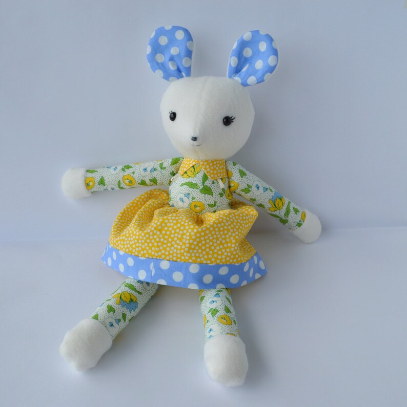 May include: A white stuffed mouse wearing a yellow and blue floral dress with white polka dots. The mouse has blue ears with white polka dots.