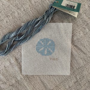 May include: A needlepoint kit featuring a square canvas with a light blue floral design. The kit includes a bundle of blue embroidery floss and a tag with product information. The canvas is white with a grid pattern.