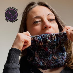 May include: A woman wearing a multi-colored knitted cowl neck scarf. The scarf has a cable knit pattern and is made with a variety of shades of blue, purple, and brown yarn.