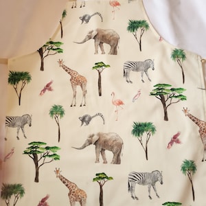 May include: A cream-coloured kitchen apron with a repeating pattern of safari animals, including giraffes, zebras, elephants, and flamingos. The animals are illustrated in a watercolour style with a light beige background.