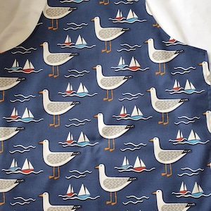 May include: A navy blue apron with a repeating pattern of seagulls, sailboats, and wavy lines. The seagulls are white and grey with orange legs and beaks. The sailboats have red, white, and blue sails. The apron has a white strap.