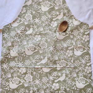 Pode incluir: Um avental verde com um padrão floral e de pássaros brancos. O avental tem um bolso grande na frente com uma colher de pau dentro.