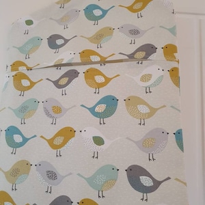 Puede incluir: Un cesto de ropa de tela blanca con un lazo para colgar en la parte superior. El cesto presenta un patrón repetitivo de pájaros coloridos con acentos amarillos, azules, grises y verdes.