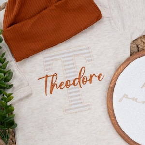 May include: A beige baby outfit set with a rust-colored ribbed knit hat. The outfit includes a long-sleeved onesie embroidered with the name "Theodore" and a decorative embroidery hoop that says "hello pumpkin".