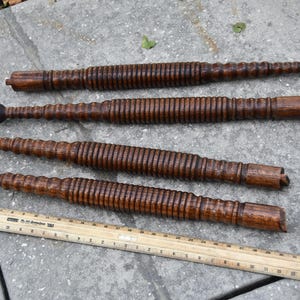 May include: Four antique wooden spindles with a dark brown finish, featuring a ribbed design and rounded ends. A wooden ruler is placed below the spindles, providing a size reference.