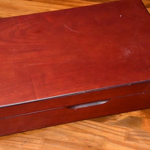 May include: A rectangular wooden storage box with a smooth, dark reddish-brown finish and visible wood grain patterns. The box features a hinged lid with a small, centered indentation along the front edge for easy opening and closing.
