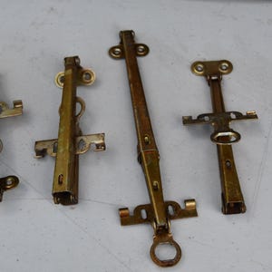 May include: Four antique brass window latches of varying sizes are arranged on a light surface. The latches have a tarnished, aged appearance, with visible signs of wear and oxidation. Each latch features a pivoting arm and mounting hardware.