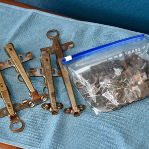 May include: Three brass-toned metal furniture hinges with articulated arms and circular mounting ends, accompanied by a clear plastic storage bag filled with a collection of small metal hardware components, including various brackets and screws.