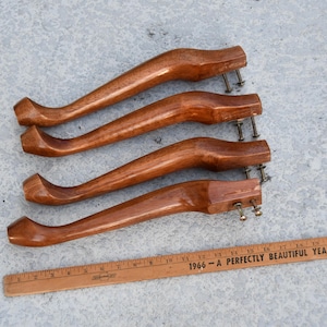 May include: Four curved, wooden furniture legs with a warm, reddish-brown finish. Each leg has metal hardware for attachment. A wooden ruler with text is in the foreground.