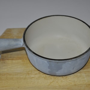 May include: A white enamel cast iron pot with a gray handle. The pot is sitting on a wooden cutting board.