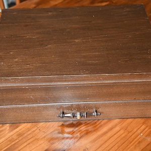 May include: A closed, rectangular wooden box with a drawer. The box is a medium brown color with visible wood grain. The drawer has a silver-colored handle. The box is sitting on a wooden surface.