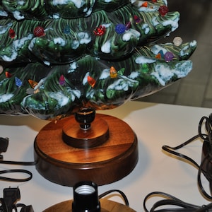 May include: A ceramic Christmas tree with green and white glaze and colorful lights on a wooden base. The tree is lit and has a cord attached to it.