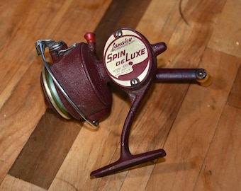 Puede incluir: Un carrete de pesca vintage de color granate con acabado metálico. La placa lateral presenta una etiqueta circular de color crema con el texto Langley Spin DeLuxe Model 830 Made in U.S.A. Incluye un arco de metal plateado y una manivela con pomo.