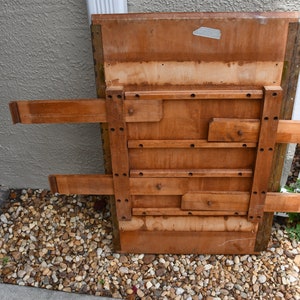 May include: The underside of a wooden table with a folding mechanism. The table is made of light brown wood and has a rectangular shape. The table is folded up and the legs are visible.