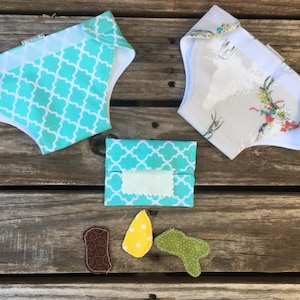 Op de afbeelding: Een verzameling handgemaakte babyartikelen, waaronder twee stoffen luiers met patronen, een bijpassende doos voor vochtige doekjes en drie decoratieve applicaties. De luiers hebben een turquoise en wit vierpaspatroon en een bloemenprint. De applicaties zijn bruin, geel en groen.