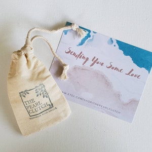 May include: A small, beige drawstring bag with the text "THE PEARL CLUTCH" and a leaf design. A card with watercolor-style blue and pink accents reads "Sending You Some Love."