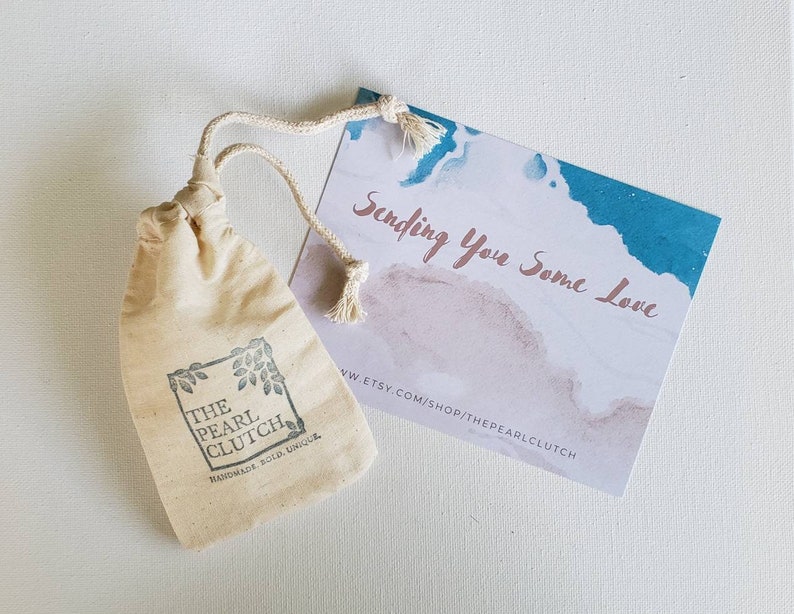 May include: A small, beige drawstring bag with the text "THE PEARL CLUTCH" and a leaf design. A card with watercolor-style blue and pink accents reads "Sending You Some Love."