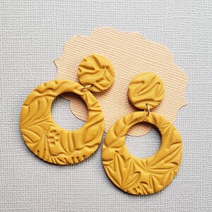 May include: Mustard yellow polymer clay earrings. Each earring features a circular design with a floral pattern and a smaller, round stud. The earrings are suspended from gold-colored hooks. The earrings are displayed on a light pink and gray background.