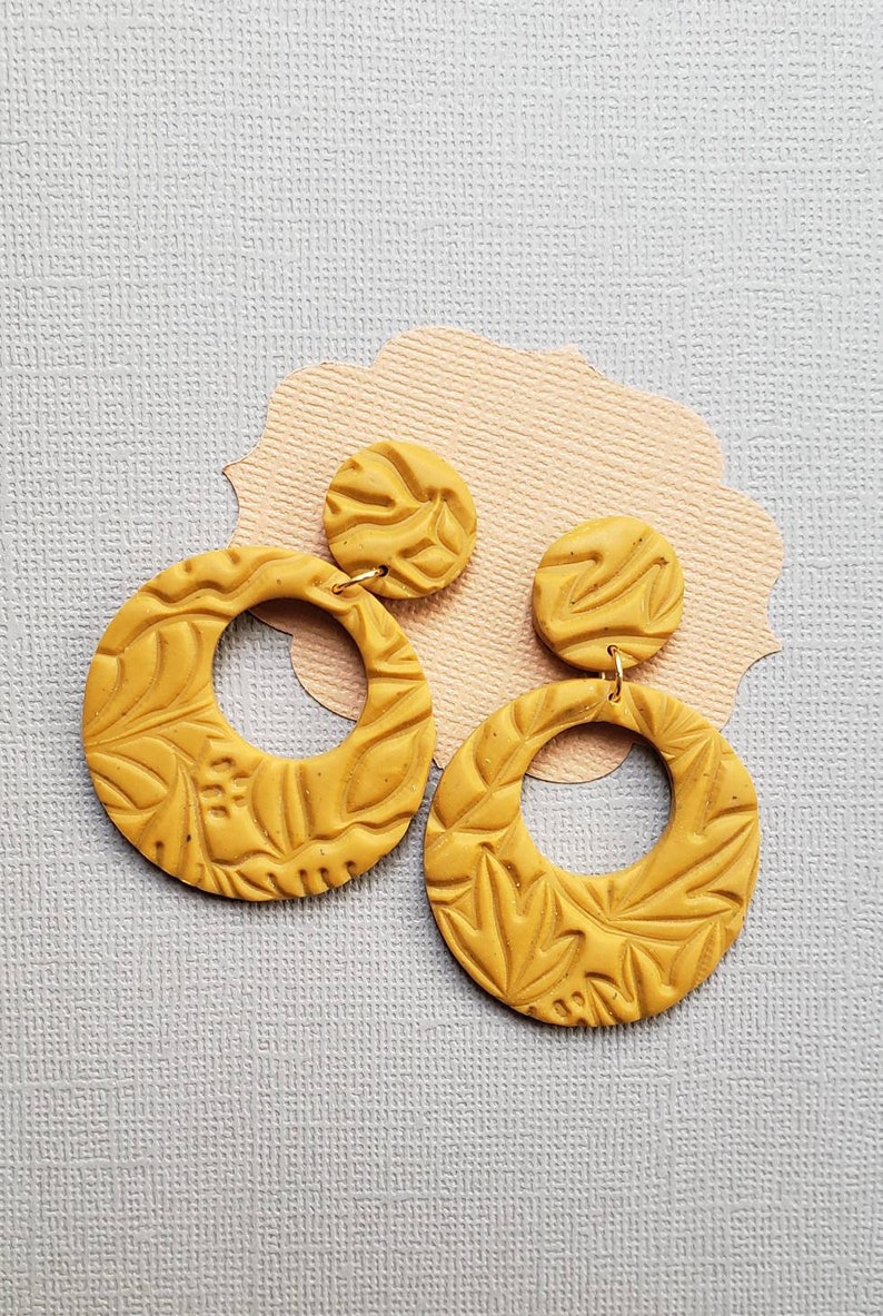 May include: Mustard yellow polymer clay earrings. Each earring features a circular design with a floral pattern and a smaller, round stud. The earrings are suspended from gold-colored hooks. The earrings are displayed on a light pink and gray background.