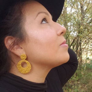 May include: Mustard yellow, textured, round earrings with a floral design. The earrings are hanging from a person's ear. The person is wearing a black turtleneck and a black hat. The background is a forest.