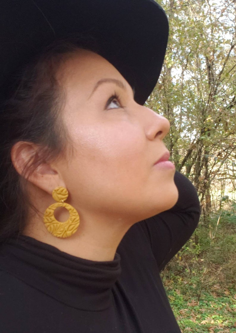 May include: Mustard yellow, textured, round earrings with a floral design. The earrings are hanging from a person's ear. The person is wearing a black turtleneck and a black hat. The background is a forest.