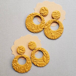 May include: Mustard yellow earrings with a floral pattern. The earrings have a circular design with a smaller, round stud at the top. The earrings are displayed on a light gray surface with a peach-colored paper cutout.