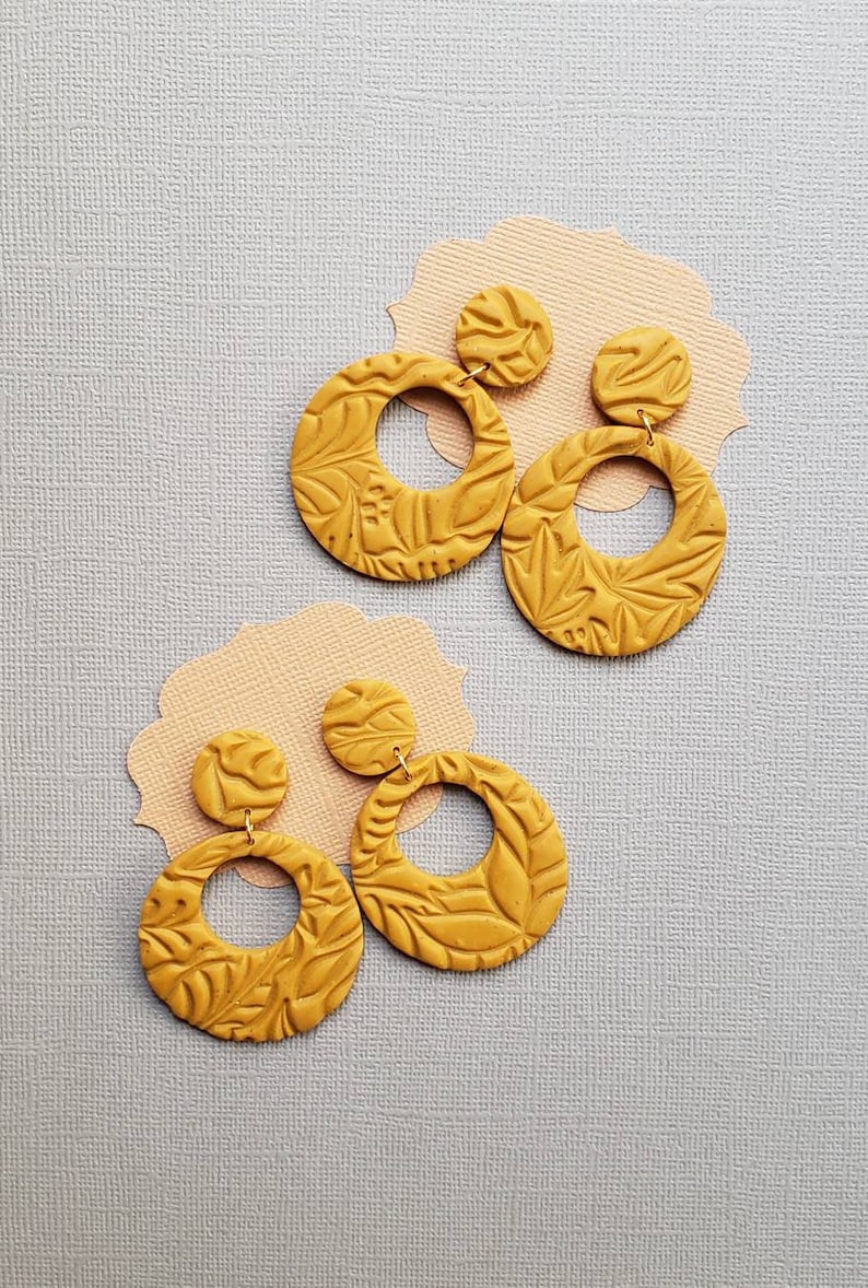 May include: Mustard yellow earrings with a floral pattern. The earrings have a circular design with a smaller, round stud at the top. The earrings are displayed on a light gray surface with a peach-colored paper cutout.