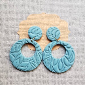 May include: A pair of light blue, textured clay earrings. Each earring features a circular design with a smaller, round stud. The earrings have a leaf-like pattern and are connected with gold-colored hooks. The earrings are displayed on a light peach-colored card.