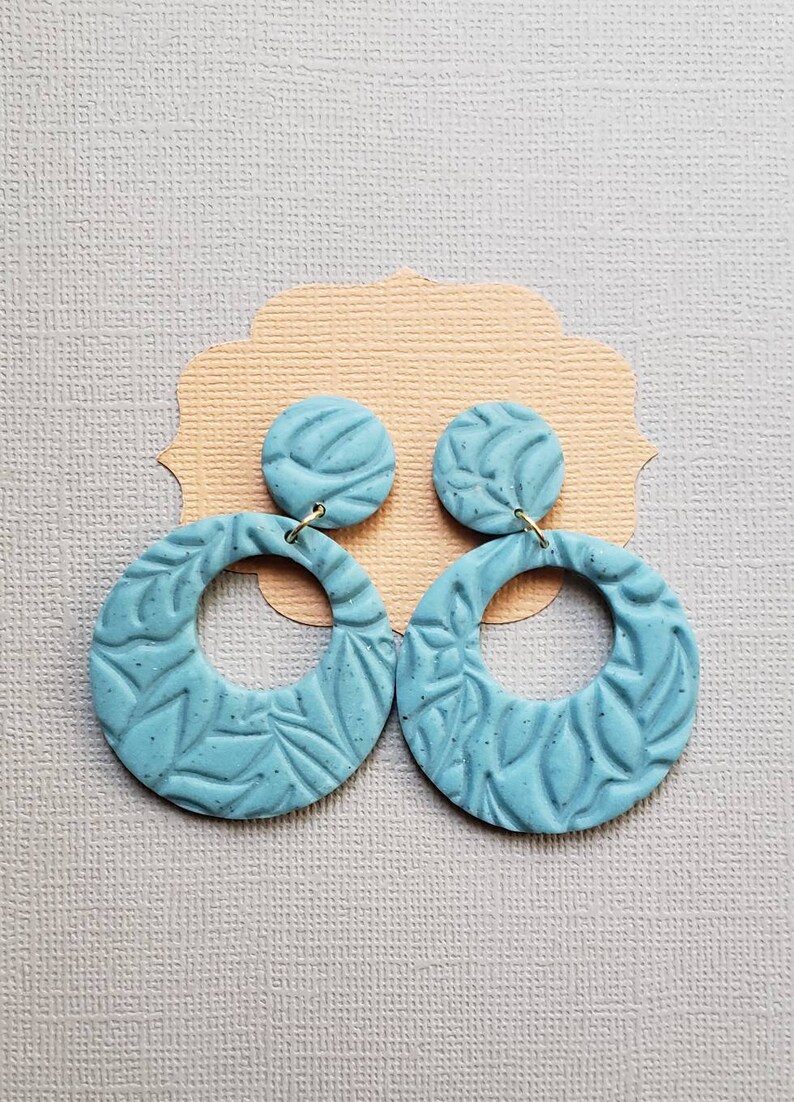 May include: A pair of light blue, textured clay earrings. Each earring features a circular design with a smaller, round stud. The earrings have a leaf-like pattern and are connected with gold-colored hooks. The earrings are displayed on a light peach-colored card.