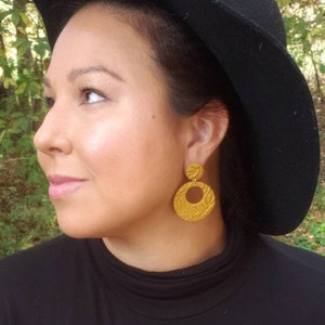 May include: Mustard yellow, textured earrings with a circular design. The earrings are worn with a black hat and black turtleneck. The earrings have a floral pattern and are a statement piece.