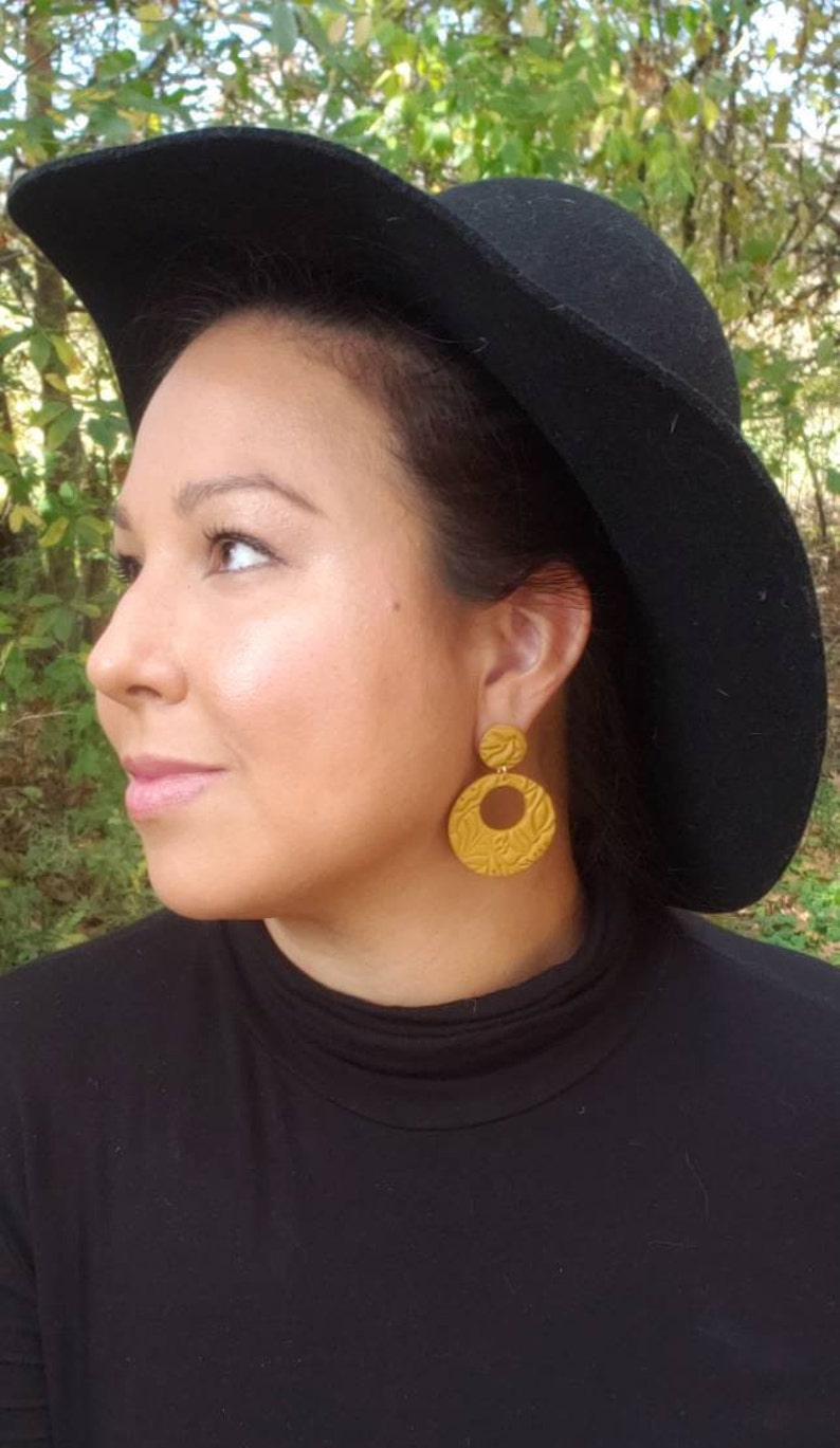 May include: Mustard yellow, textured earrings with a circular design. The earrings are worn with a black hat and black turtleneck. The earrings have a floral pattern and are a statement piece.