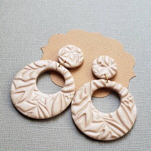 May include: A pair of beige, textured earrings. Each earring features a circular design with a floral pattern. The earrings are attached to small, round studs with a similar design. The earrings are displayed on a brown, decorative paper cutout.